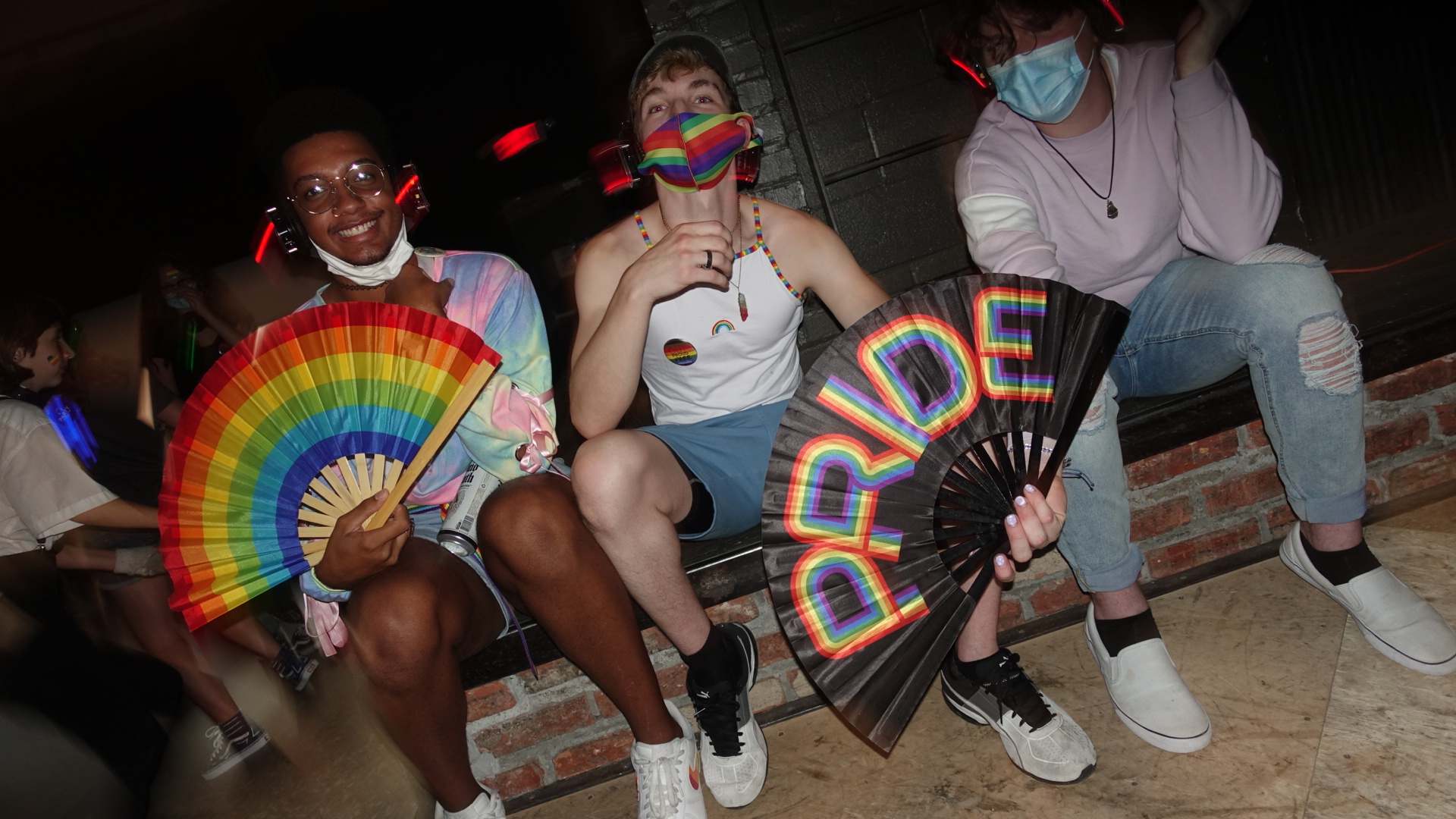 Teens in rainbow masks hold rainbow fans with rainbow pins. The background is blurry and distorted, as if taken in motion.