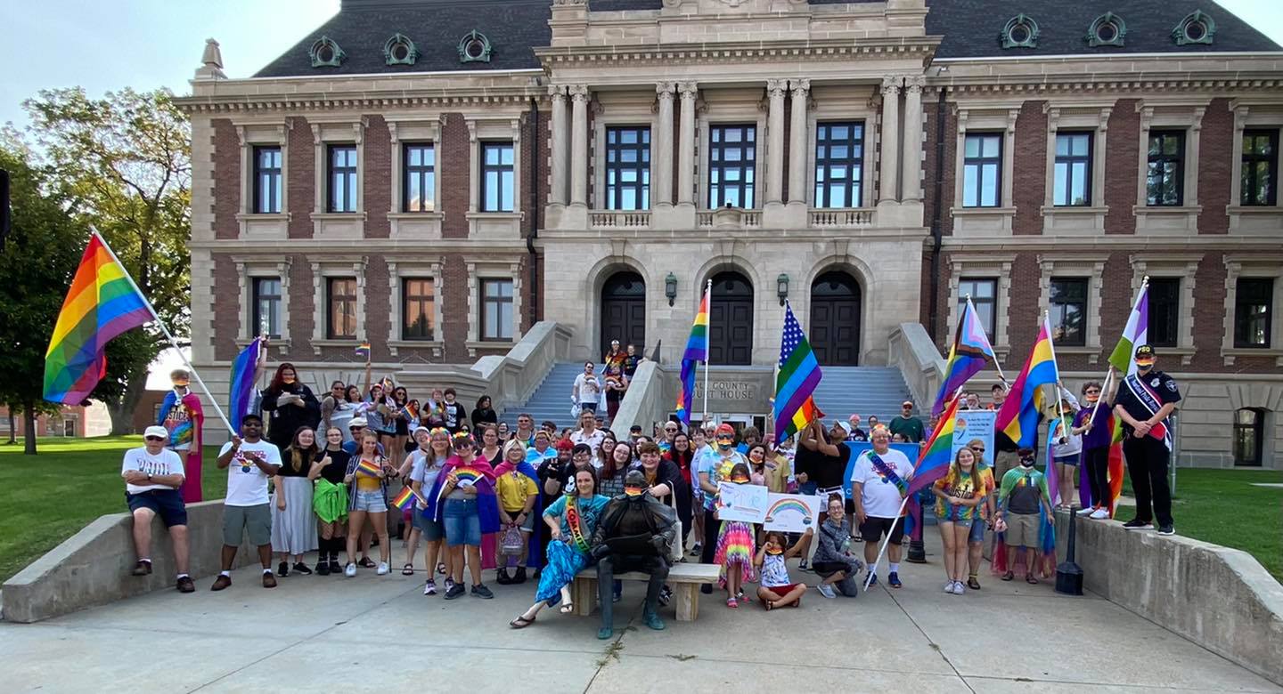 A large group of people wearing rainbow colors, pride flag capes, and holding rainbow flags, stand in front of the Hall County Court House.