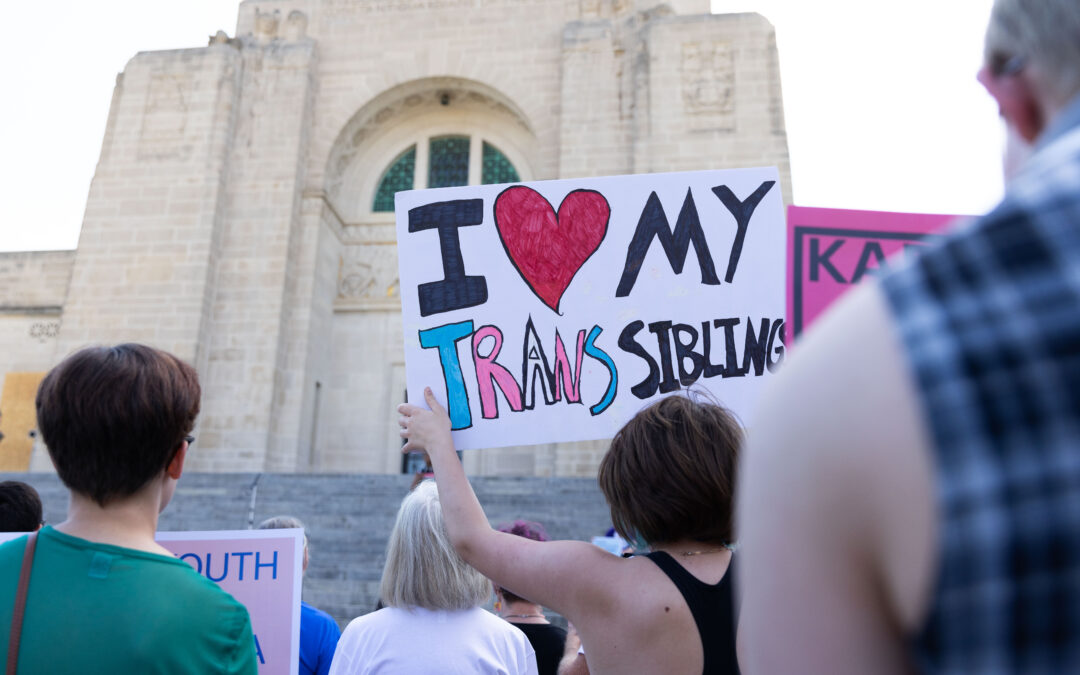 Photo of a rally attendee holding a sign that reads "I love my trans sibling" on the steps of the Nebraska capitol.