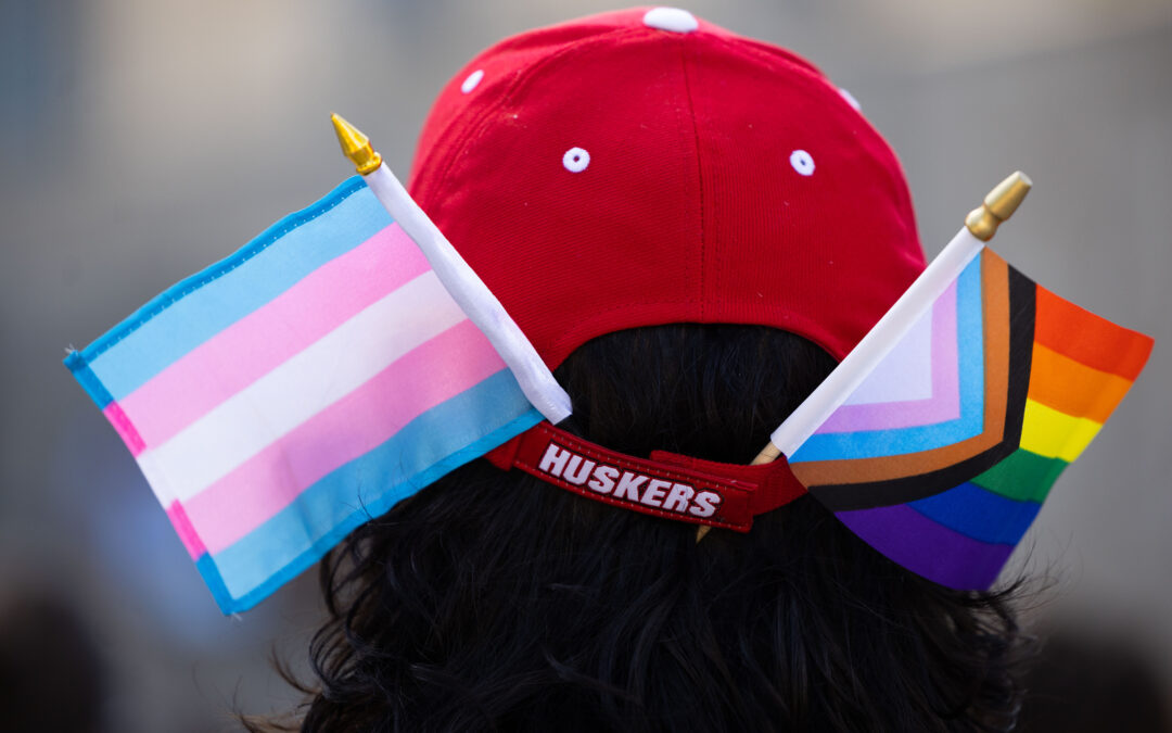 Photo of someone wearing a red baseball cap that reads "Huskers" on the back strap. They have a mini trans flag and a mini progress flag tucked into the back of the hat on either side.