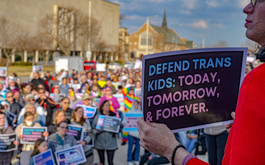 A person stands in front of a crowd at the Trans Day of Visibility rally. They are holding a sign that says "Defend trans youth: today, tomorrow and forever."