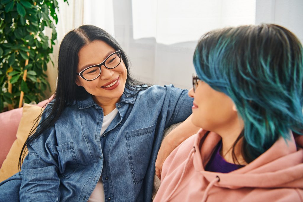 Parent and teenage child engage in heartfelt conversation on a cozy couch at home, creating a warm and loving atmosphere. A smiling parent in glasses talks with a blue-haired teen in a pink hoodie on the couch, showing how families support transgender youth.